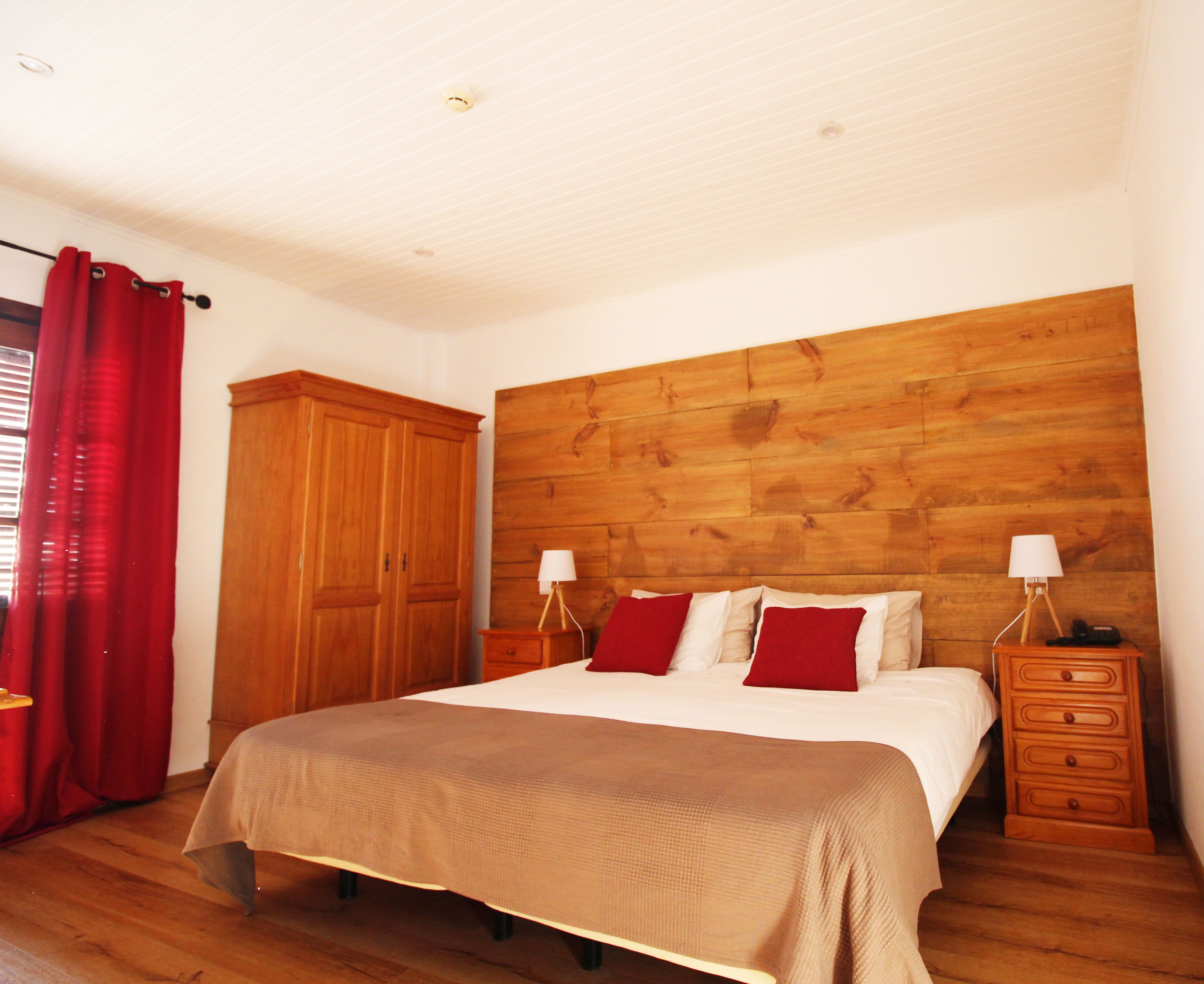 Une chambre à coucher avec une tête de lit en bois, un lit recouvert d’une couvre-lit beige, deux tables de chevet avec des lampes, une armoire en bois et des rideaux rouges.