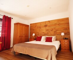 Une chambre à coucher avec une tête de lit en bois, un lit recouvert d’une couvre-lit beige, deux tables de chevet avec des lampes, une armoire en bois et des rideaux rouges.