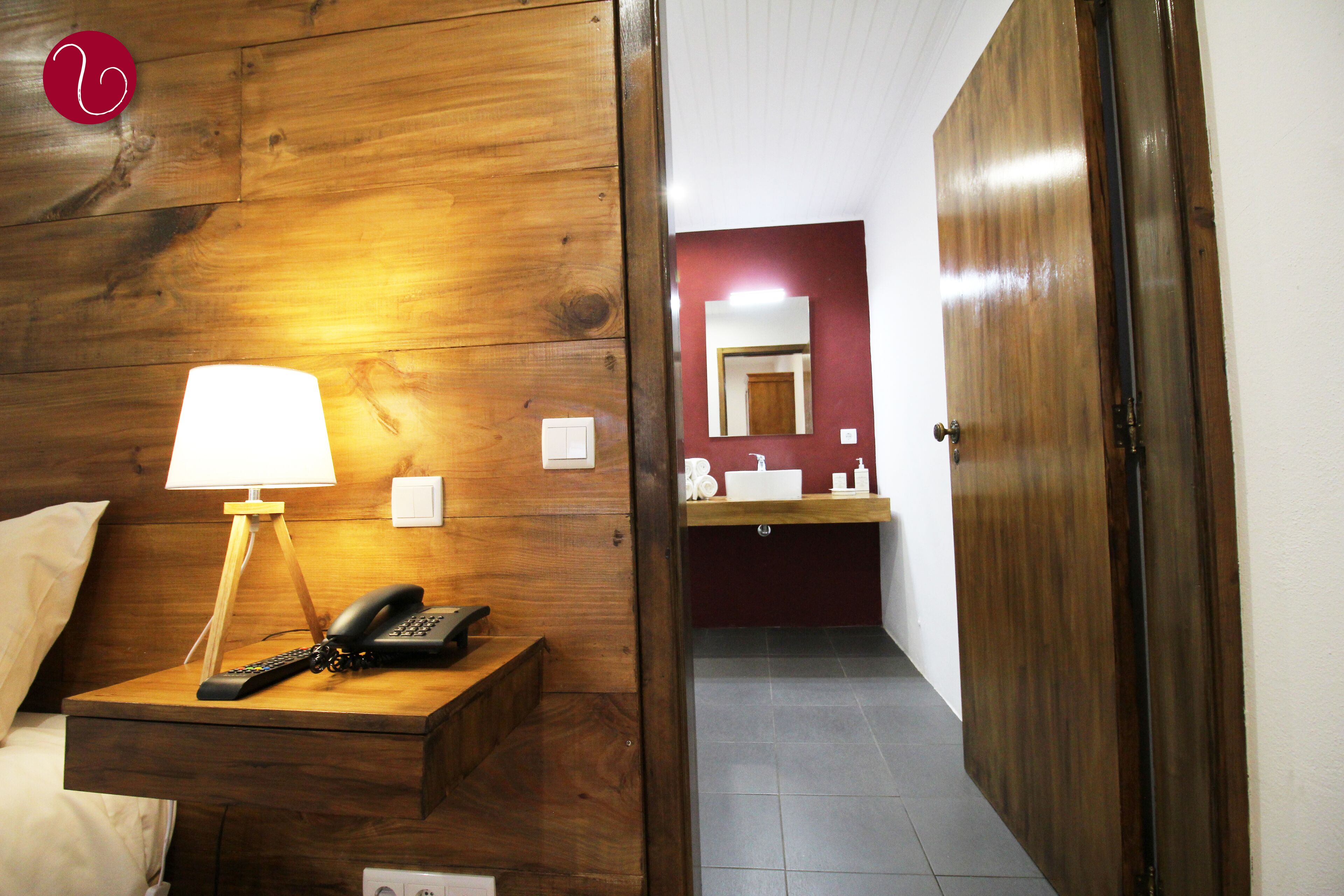 A hotel room with a wooden headboard, a bedside lamp, a telephone, a mirror, and a bathroom with a sink and mirror.
