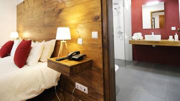Une chambre d’hôtel avec une tête de lit en bois, une table de chevet avec une lampe et un téléphone, un lit avec des oreillers rouges et une salle de bain avec un lavabo et un miroir.