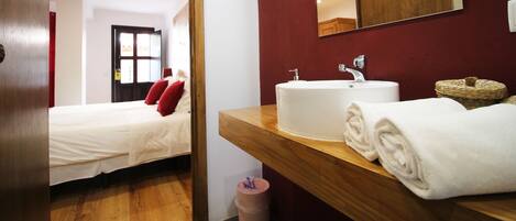 Habitación de hotel con una cama, una mesita de noche de madera, un lavamanos blanco y una toalla, y una pared roja con un espejo.