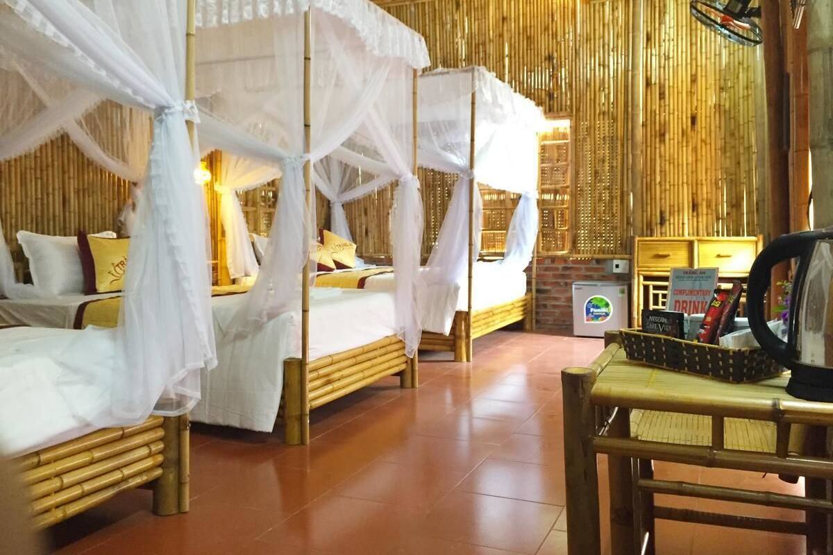 A room with four beds, each with a canopy. There is a small table with a kettle and a basket of snacks. The room has a bamboo wall and a tiled floor.