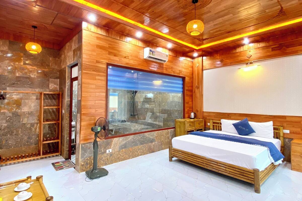 A room with a bed, wooden furniture, a fan, a table with teapots, and a stone wall.