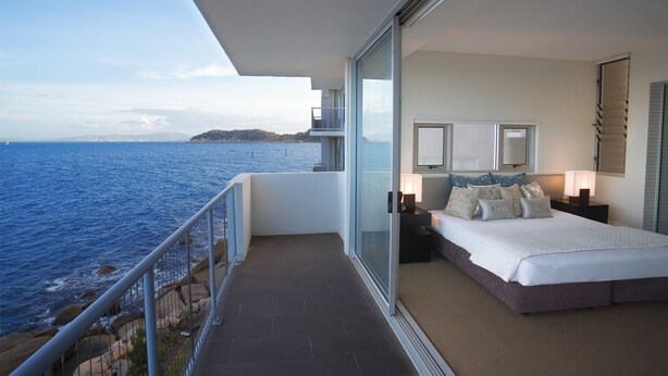 A balcony with a view of the sea, a bed with a white duvet and multiple pillows, and a nightstand with a lamp. - 1 Bright Point Apartment 5201 (Nelly Bay)