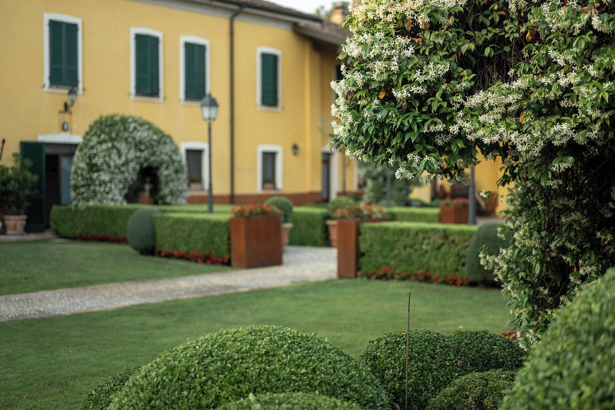Un jardín bien cuidado con setos ordenados y un sendero que conduce a un edificio amarillo con persianas verdes.