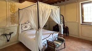 Una cama con dosel sobre cuatro postes, con estructura de madera y ropa de cama blanca, una pequeña silla de madera y piso de baldosas.