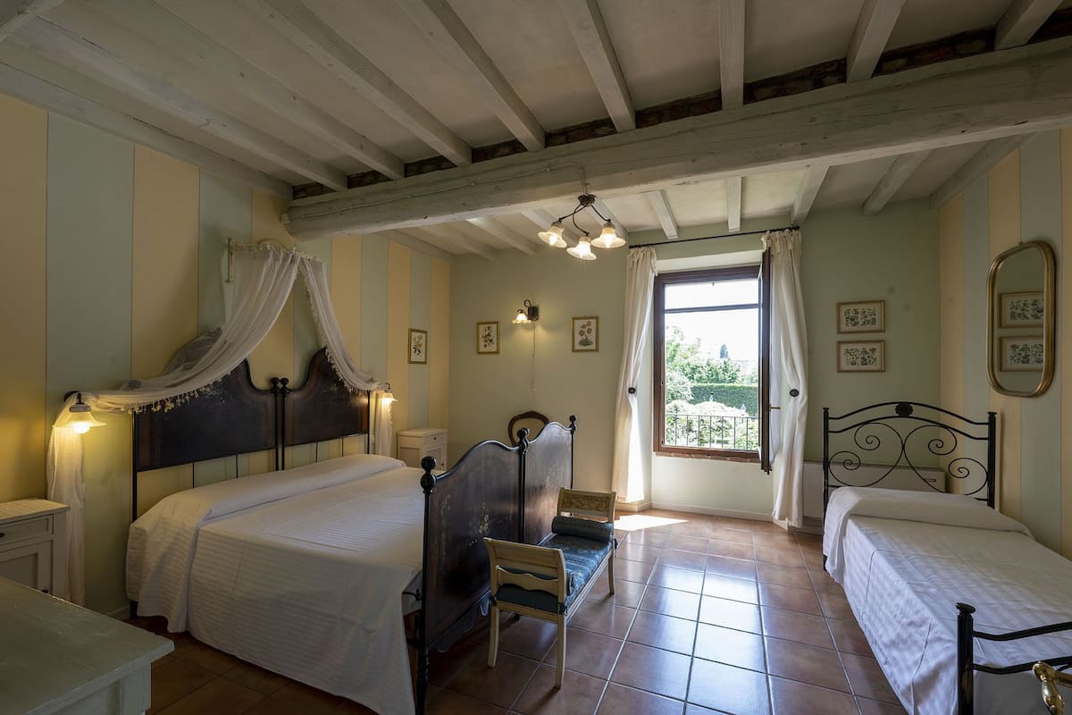 A bedroom with two beds, a wooden bench, a nightstand, and a window with curtains.