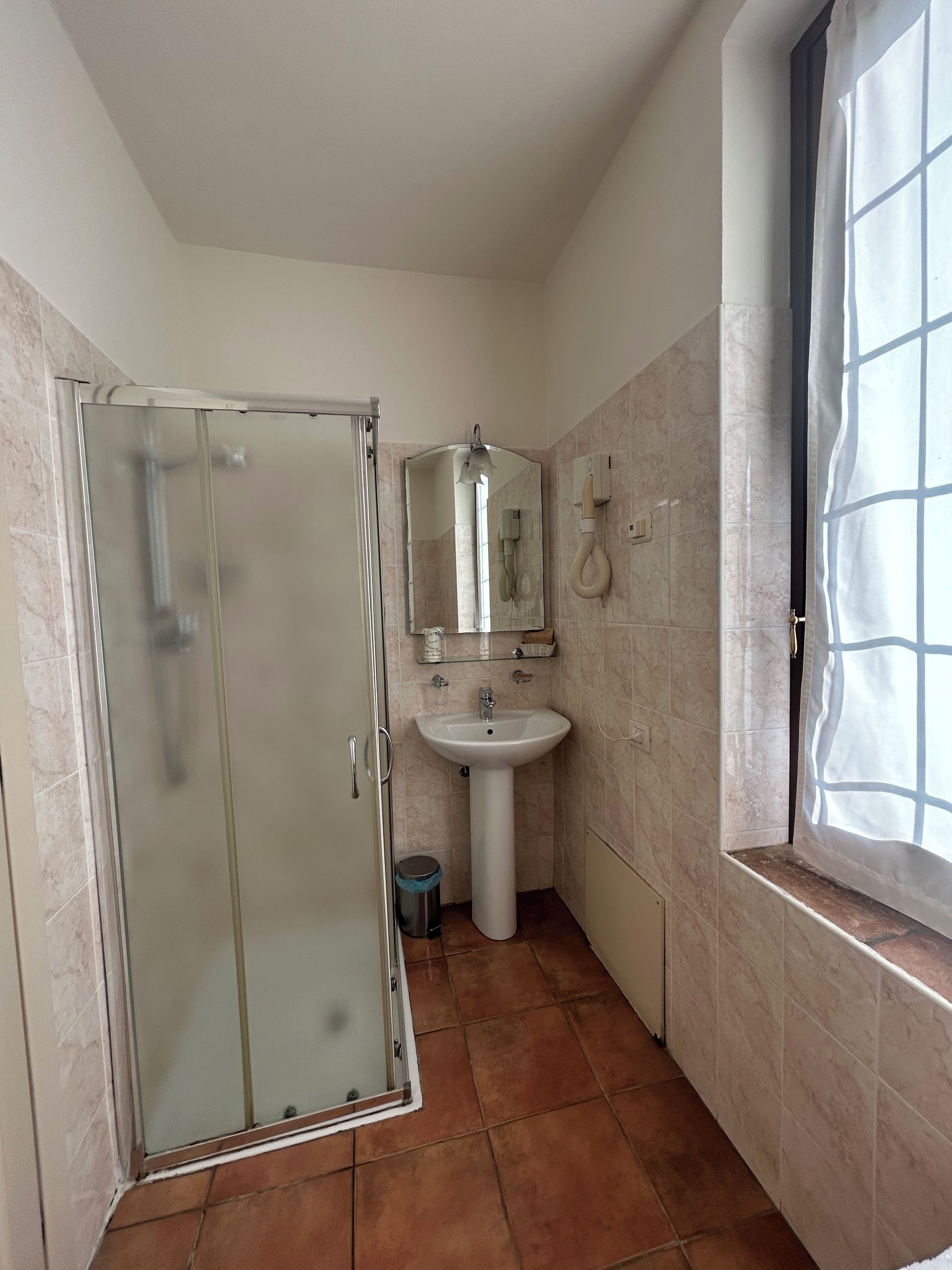 Un baño con cabina de ducha de vidrio, lavamanos sobre pedestal y ventana con cortina.