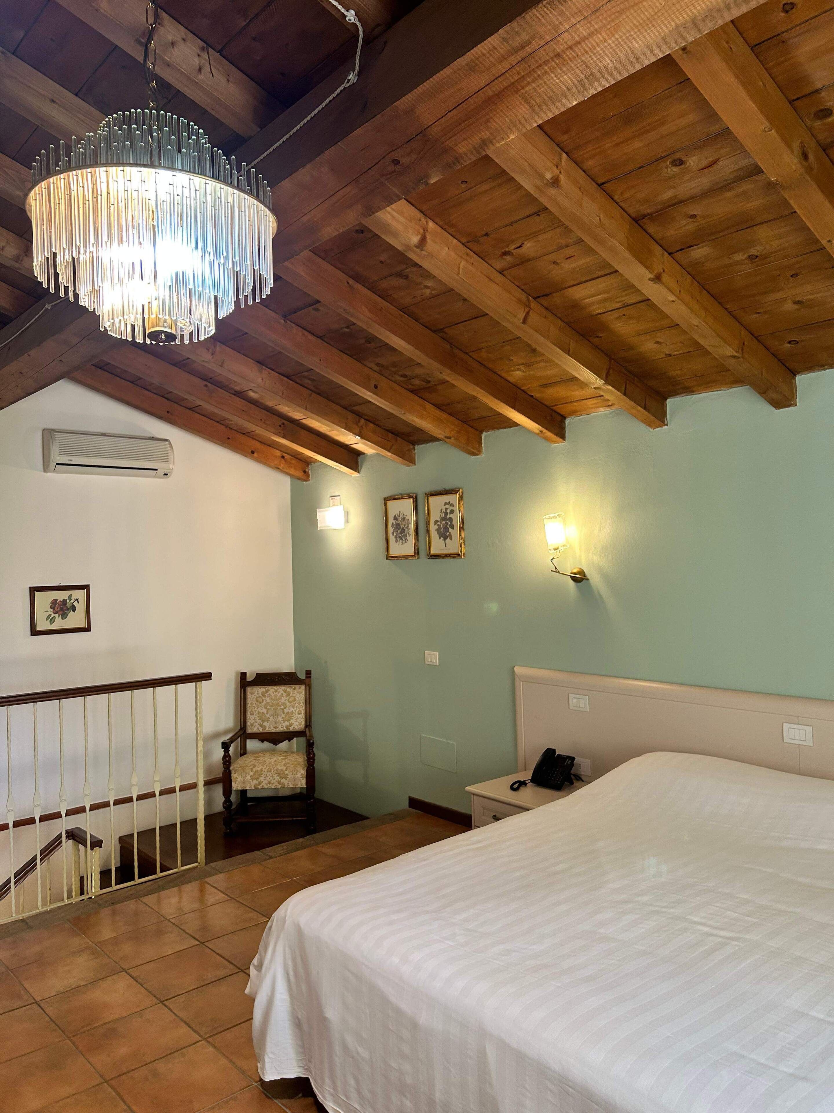 A bedroom with a wooden ceiling, a bed, a chair, a nightstand, and a wall-mounted light fixture.