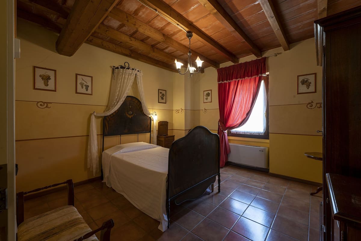 A bedroom with a wooden ceiling, a bed with a canopy, a nightstand, a chair, and a window with red curtains.