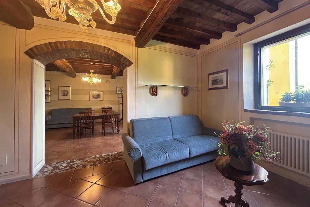 A living room with a blue sofa, a dining table with chairs, a chandelier, and a window with plants.