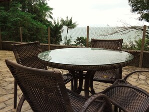A round glass table surrounded by wicker chairs on a patio with a view of the sea.
