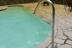 A swimming pool with a metal ladder, stone flooring, and a stone wall in the background.
