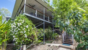 A modern two-story house with a balcony, surrounded by lush greenery and a variety of plants.