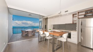 Een moderne keuken met een groot eiland, witte stoelen en uitzicht op de zee via een open balkon.