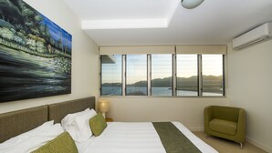A hotel room with a large bed, a green armchair, a painting of a landscape, and a view of a body of water through large windows. - 1 Bright Point Apartment 4201 (Nelly Bay)