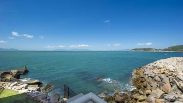 A coastal view with a rocky breakwater, clear blue sea, and distant mountains.