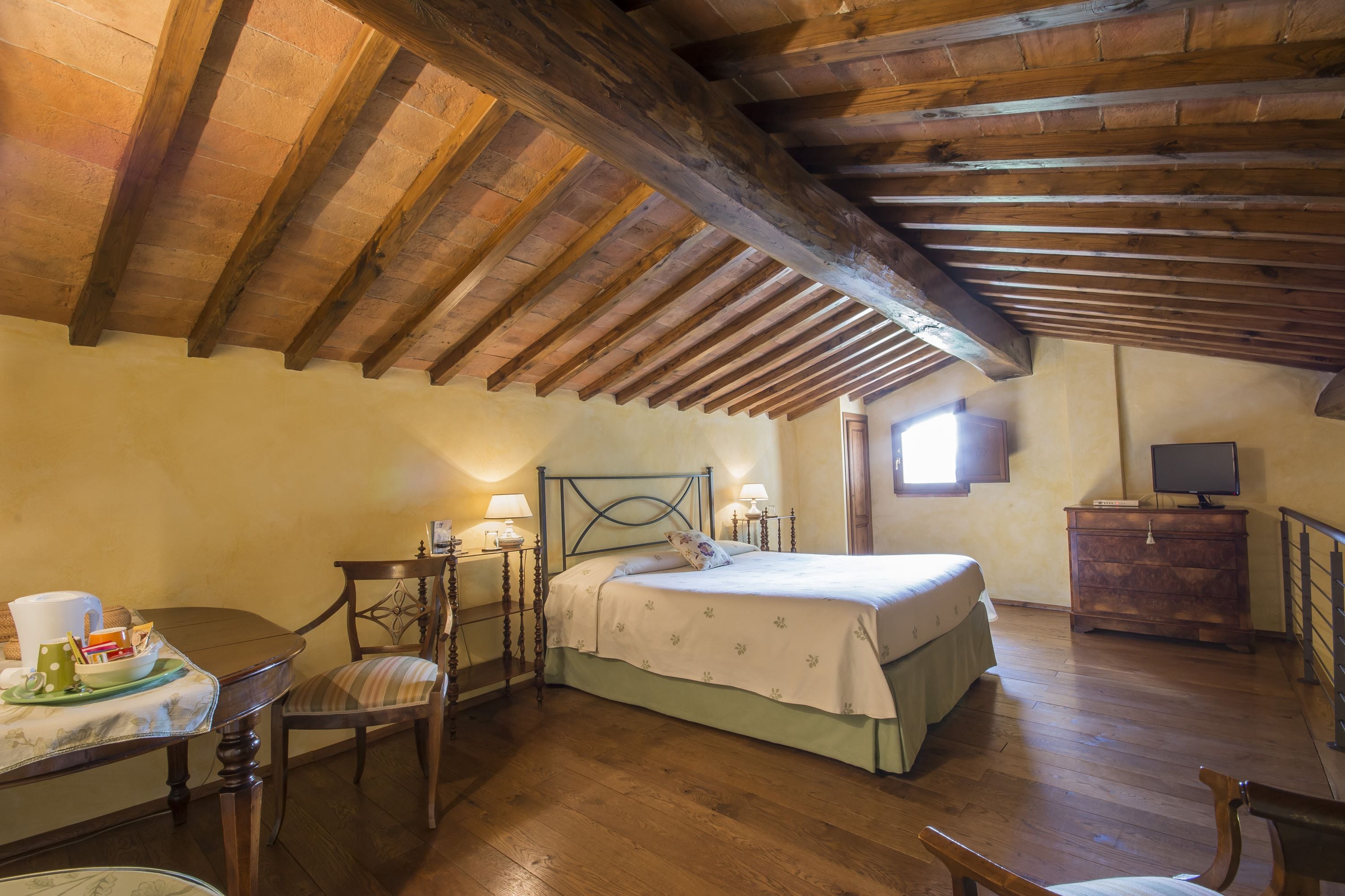 A bedroom with a wooden ceiling, a bed with a green bedspread, a bedside table with a lamp, a chair, and a dresser with a TV.