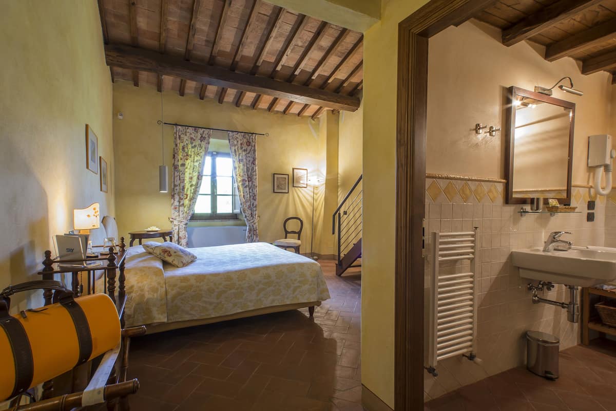 Un dormitorio con cama, escritorio con computadora portátil, silla, ventana con cortinas y baño con lavamanos y espejo.