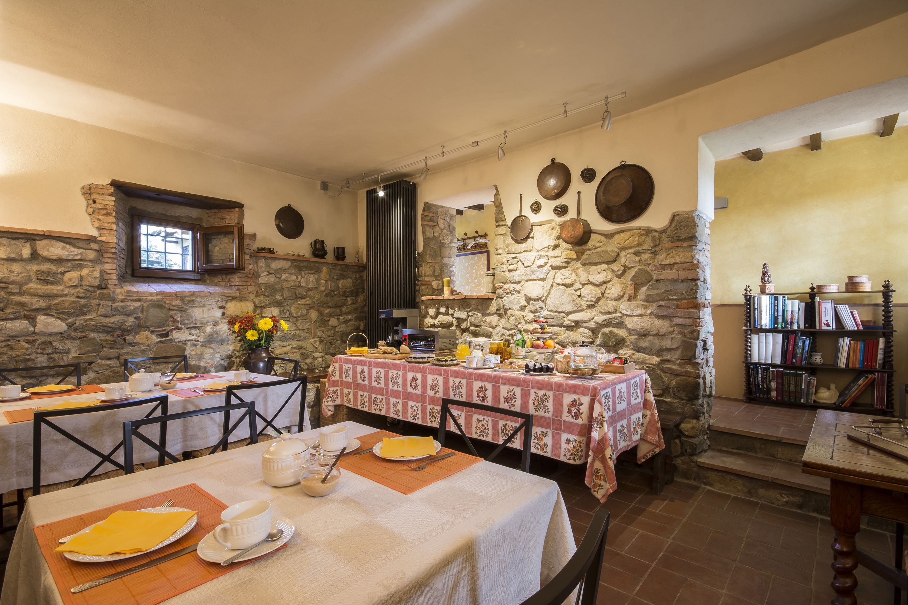 Un coin repas aux murs de pierre, avec une cheminée en pierre et une bibliothèque. Les tables sont dressées avec des assiettes, des tasses et des couverts.