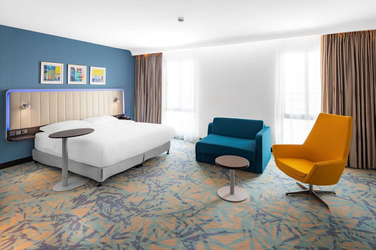 Een hotelkamer met een bed, een blauwe fauteuil, een gele fauteuil en een klein tafeltje.
