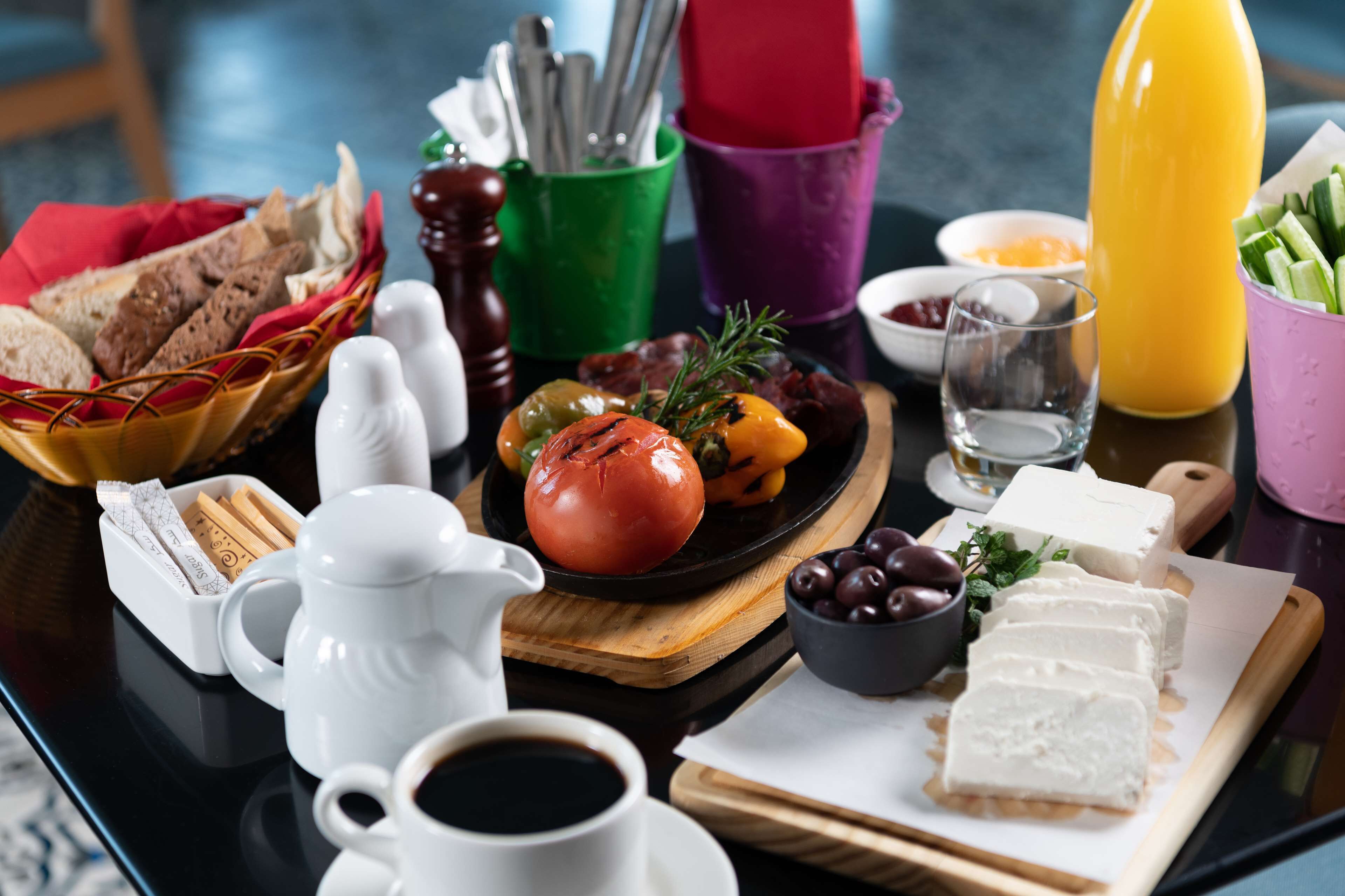 A table with a variety of food items including bread, cheese, olives, and a tomato, along with condiments and a jug of juice.