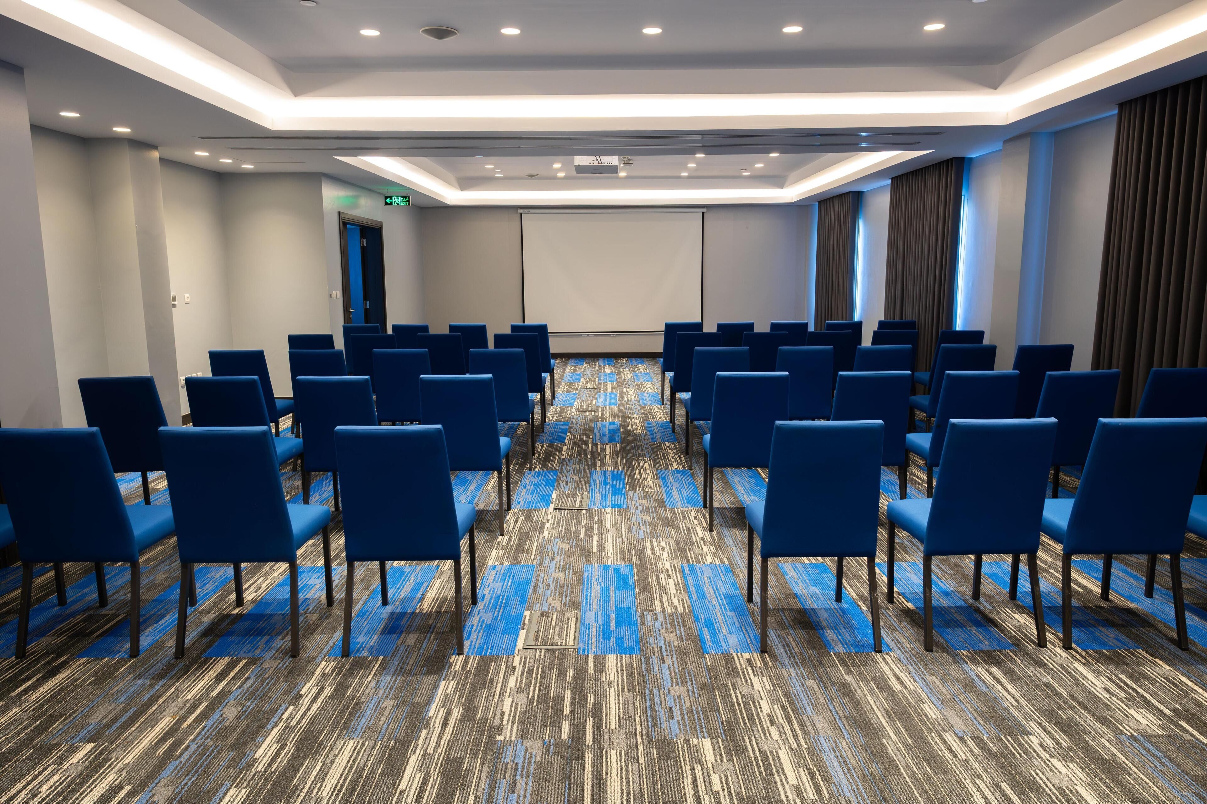 A conference room with rows of blue chairs, a patterned carpet, and a projector screen.