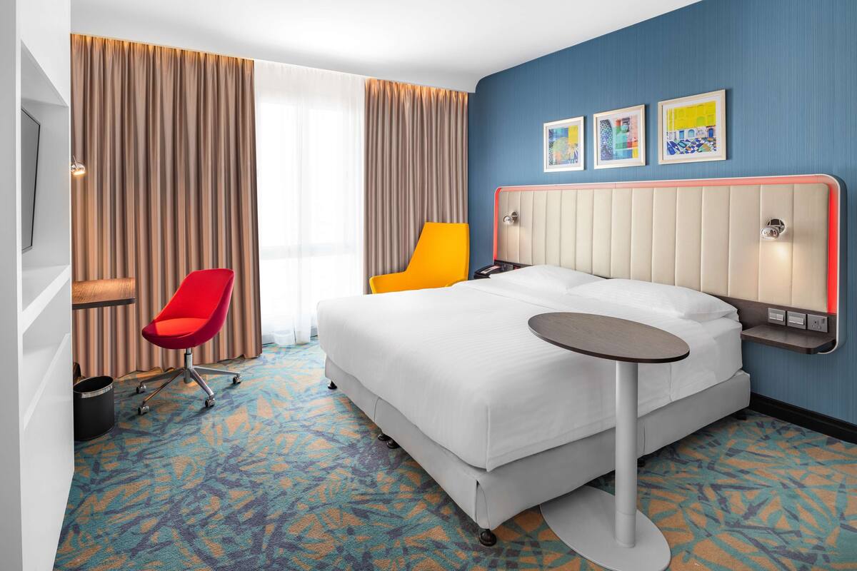 Een hotelkamer met een bed, een rode stoel, een klein tafeltje en een raam met gordijnen.