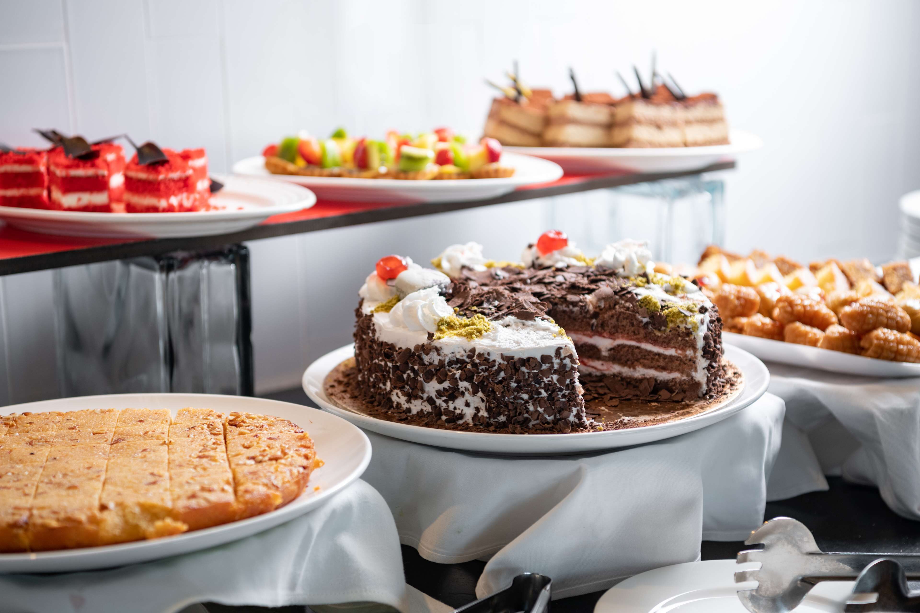 A variety of desserts on a buffet table, including a chocolate cake with whipped cream and cherries, a red velvet cake, and other assorted pastries.