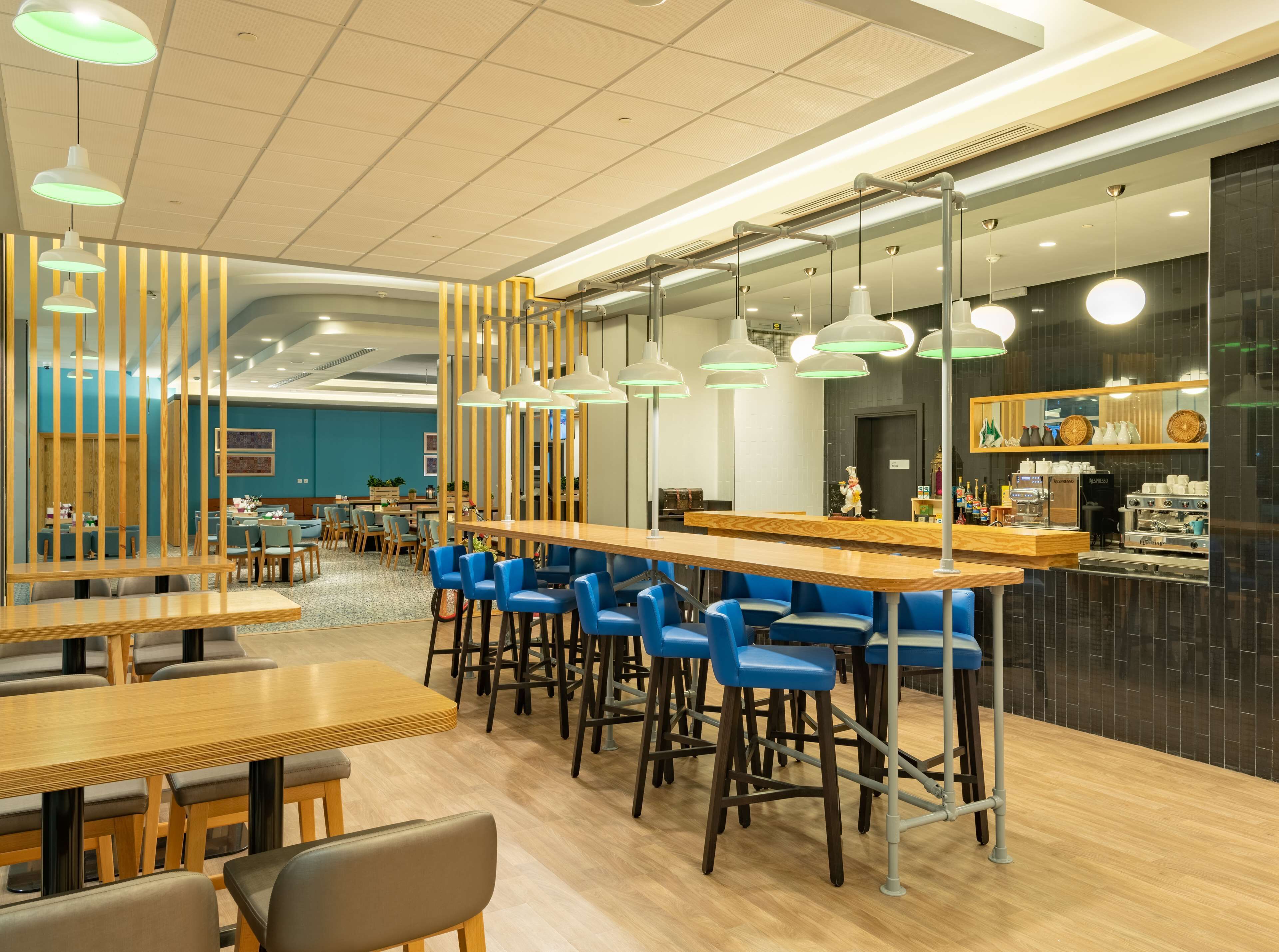Un café moderno con mesas de madera, taburetes de barra azules, una barra con máquinas de café e iluminación colgante.