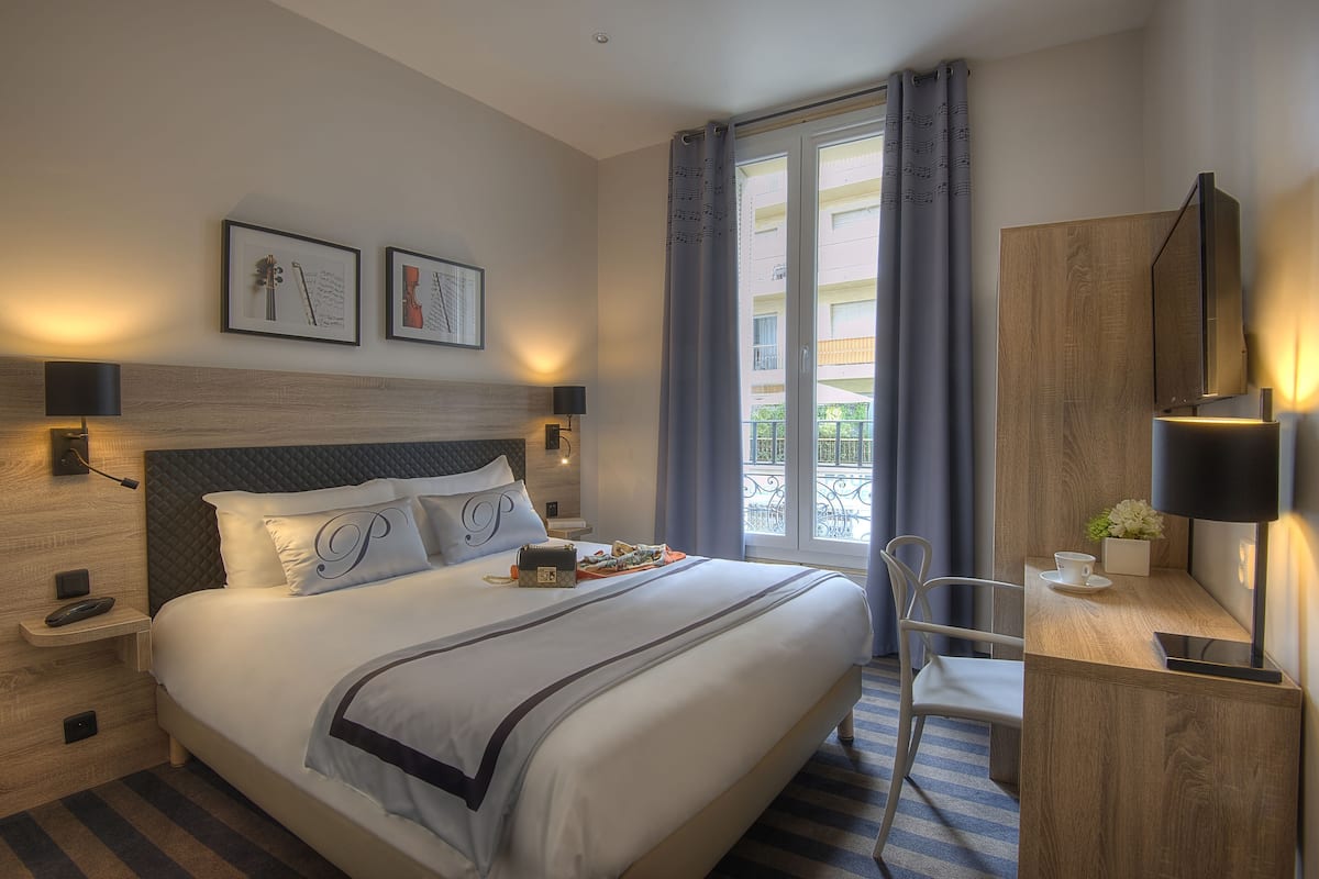 A hotel room with a large bed, bedside tables, a desk with a chair, a TV, and a window with curtains.