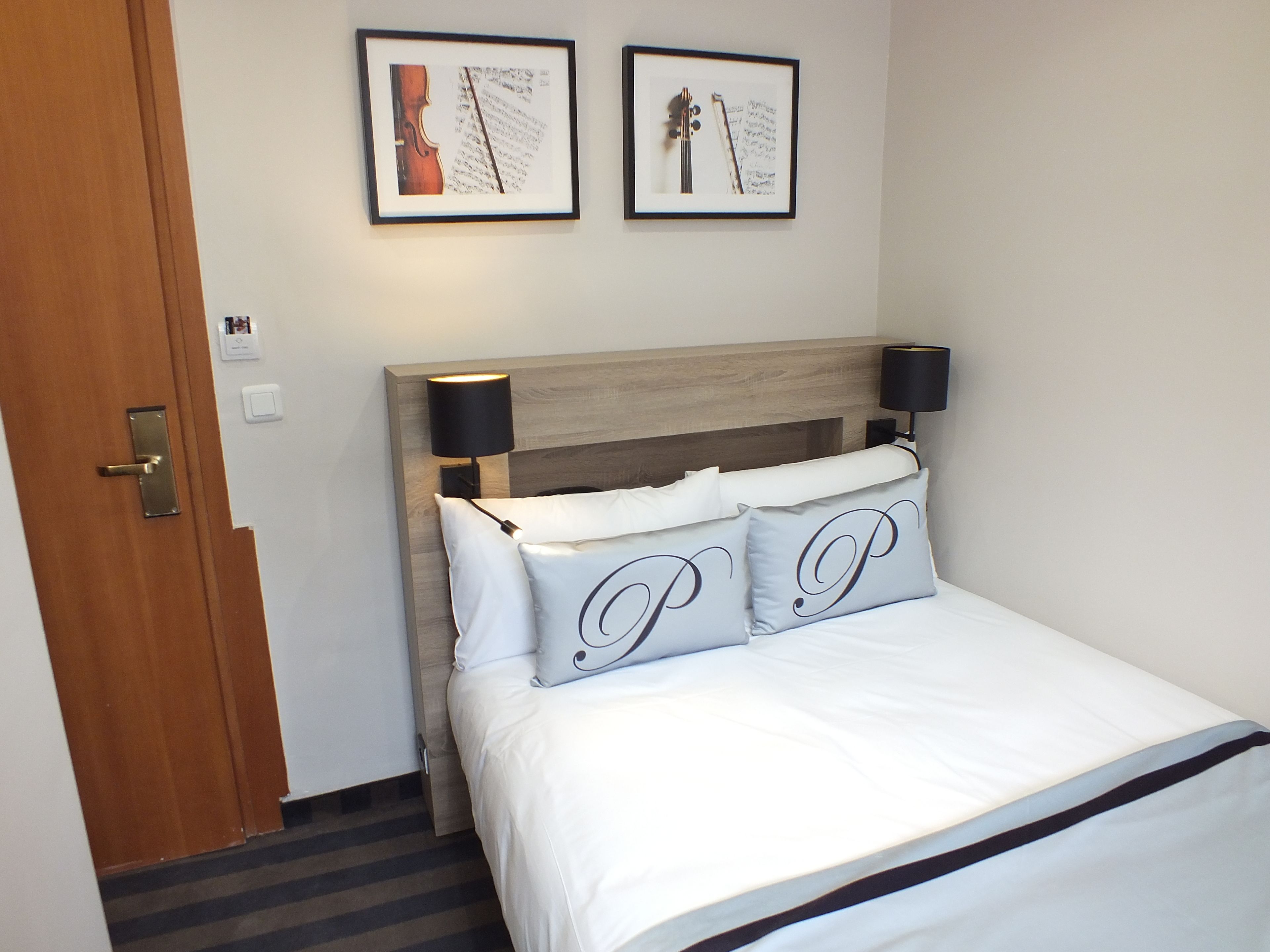 A hotel room with a wooden headboard, two bedside lamps, a bed with white bedding, and two framed pictures on the wall.