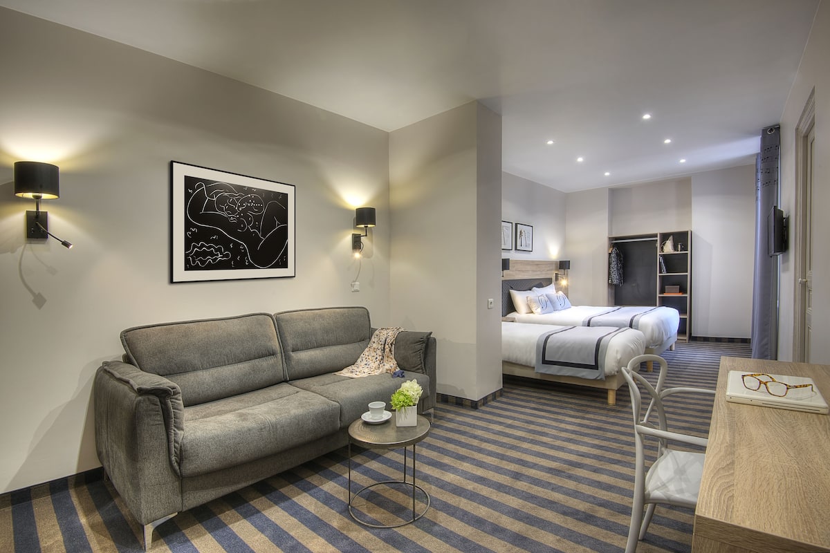 A modern hotel room with a grey sofa, a small round table with a vase, a bed with a white duvet, and a wardrobe.