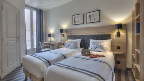 Een hotelkamer met twee bedden, een bureau en een stoel. Twee wandlampen, een boekenkast en een raam met gordijnen.