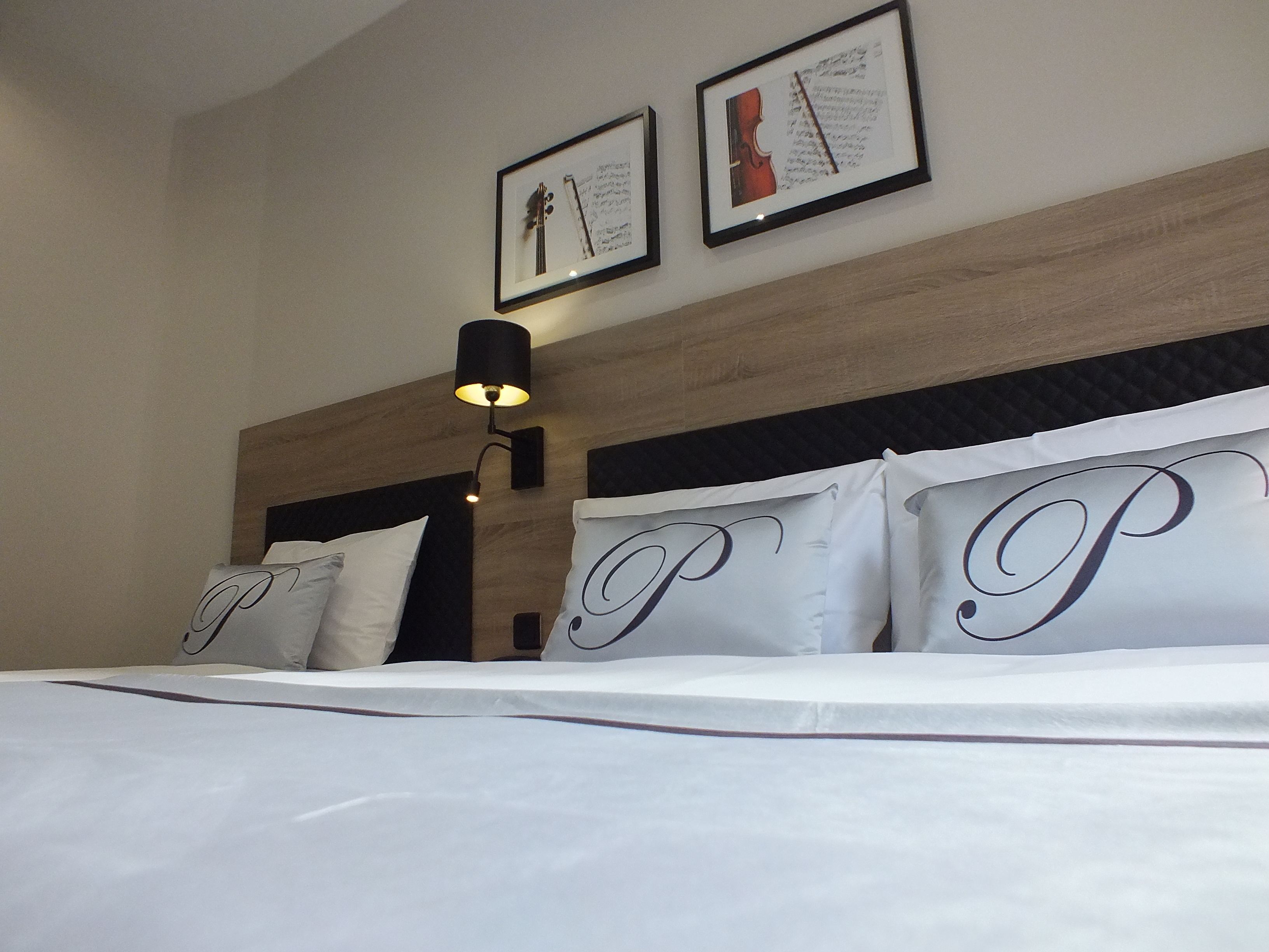 Habitación de hotel con una cama grande, cabecera con panel de madera, dos cuadros enmarcados en la pared y una lámpara fijada a la pared.