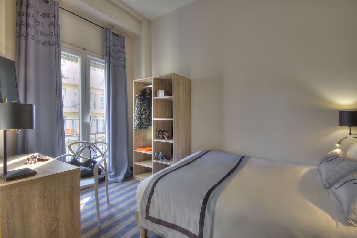 A hotel room with a bed, a desk with a lamp, a chair, a wardrobe, and a window with curtains.
