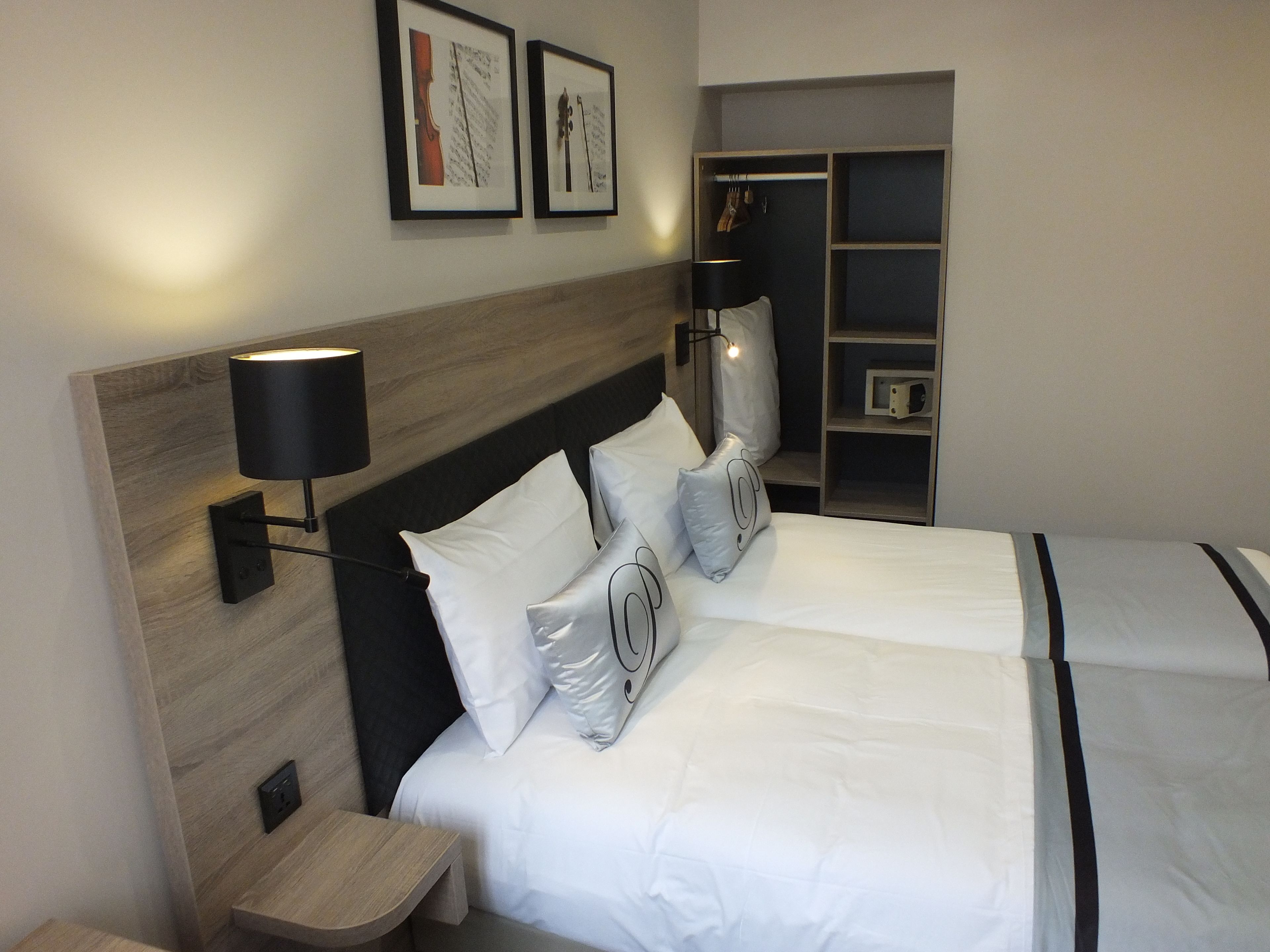 Een hotelkamer met twee bedden, een houten hoofdbord, een nachtkastje, een lamp, een kledingkast en twee ingelijste foto's aan de muur.