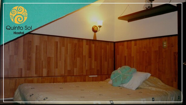 Quarto com paredes de madeira, uma cama com colcha estampada, luminária fixada na parede e uma prateleira sobre a cama.