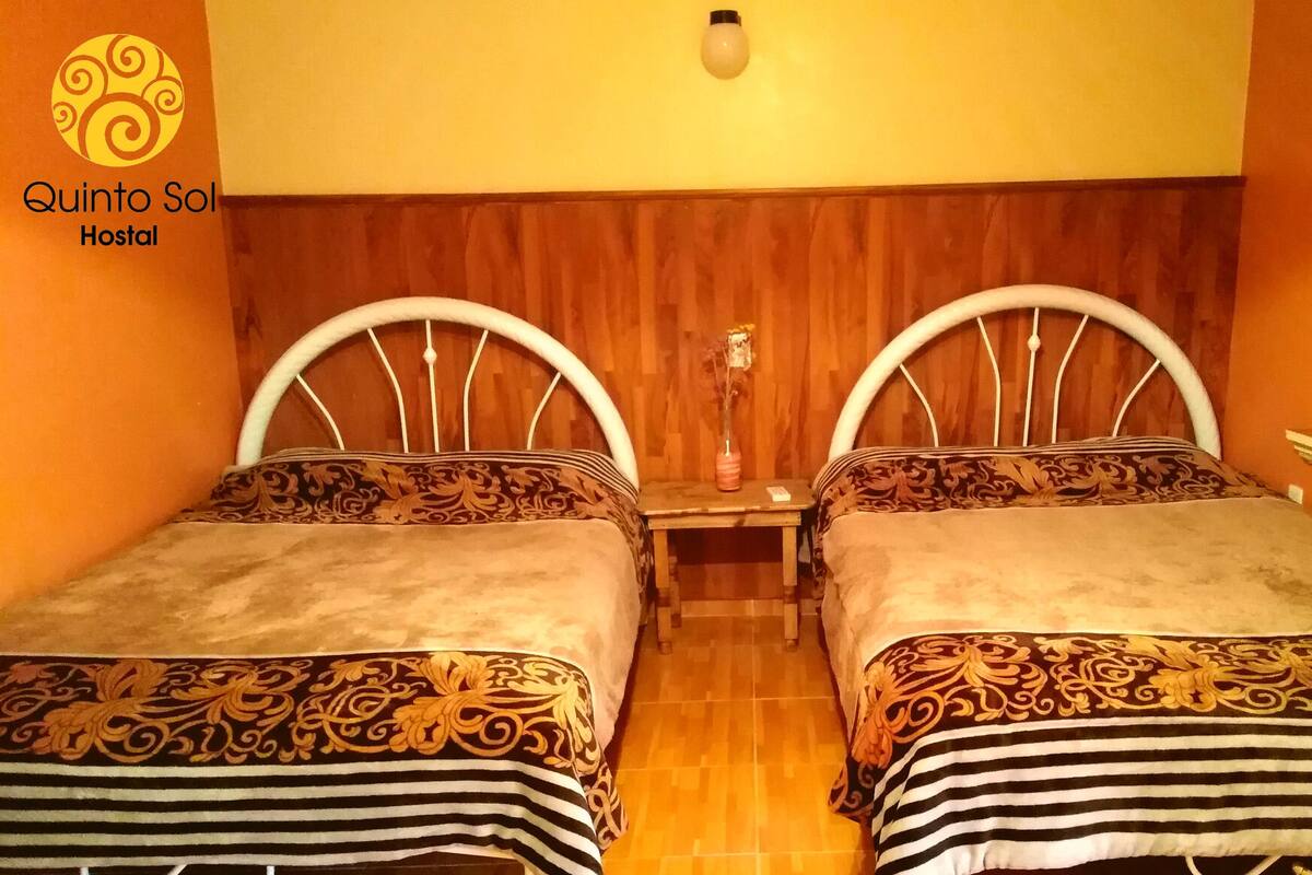 A hotel room with two beds, wooden headboards, a small table with a vase, and a mirror above the table.