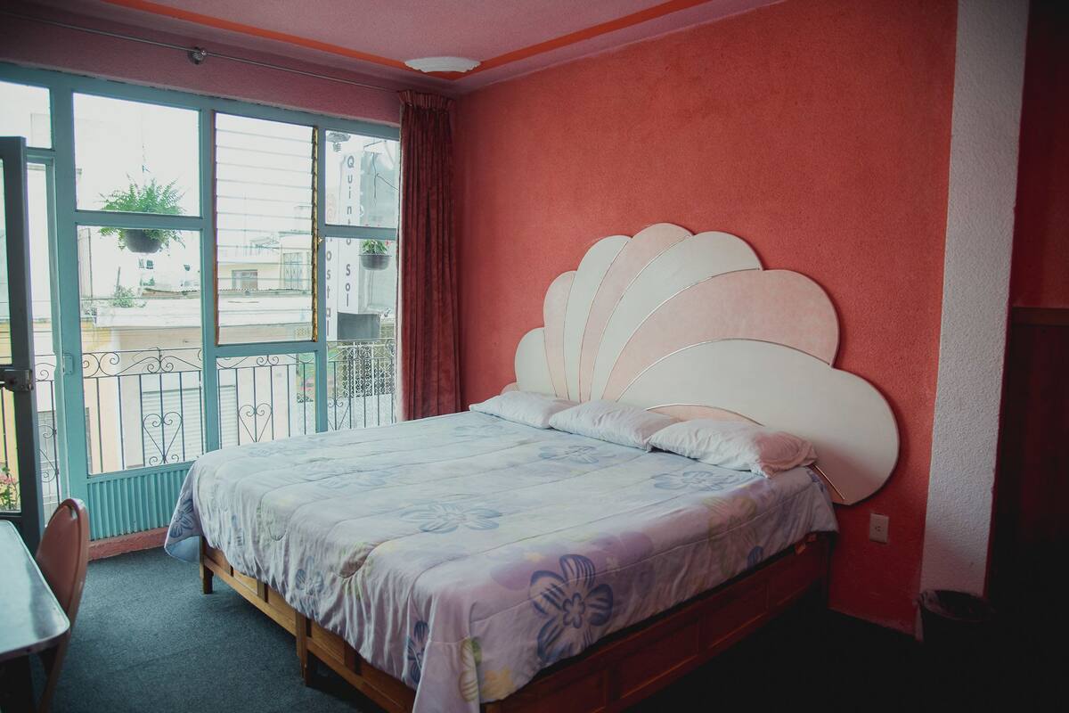 A bedroom with a large bed, a window with a view of buildings, and a balcony with a railing.