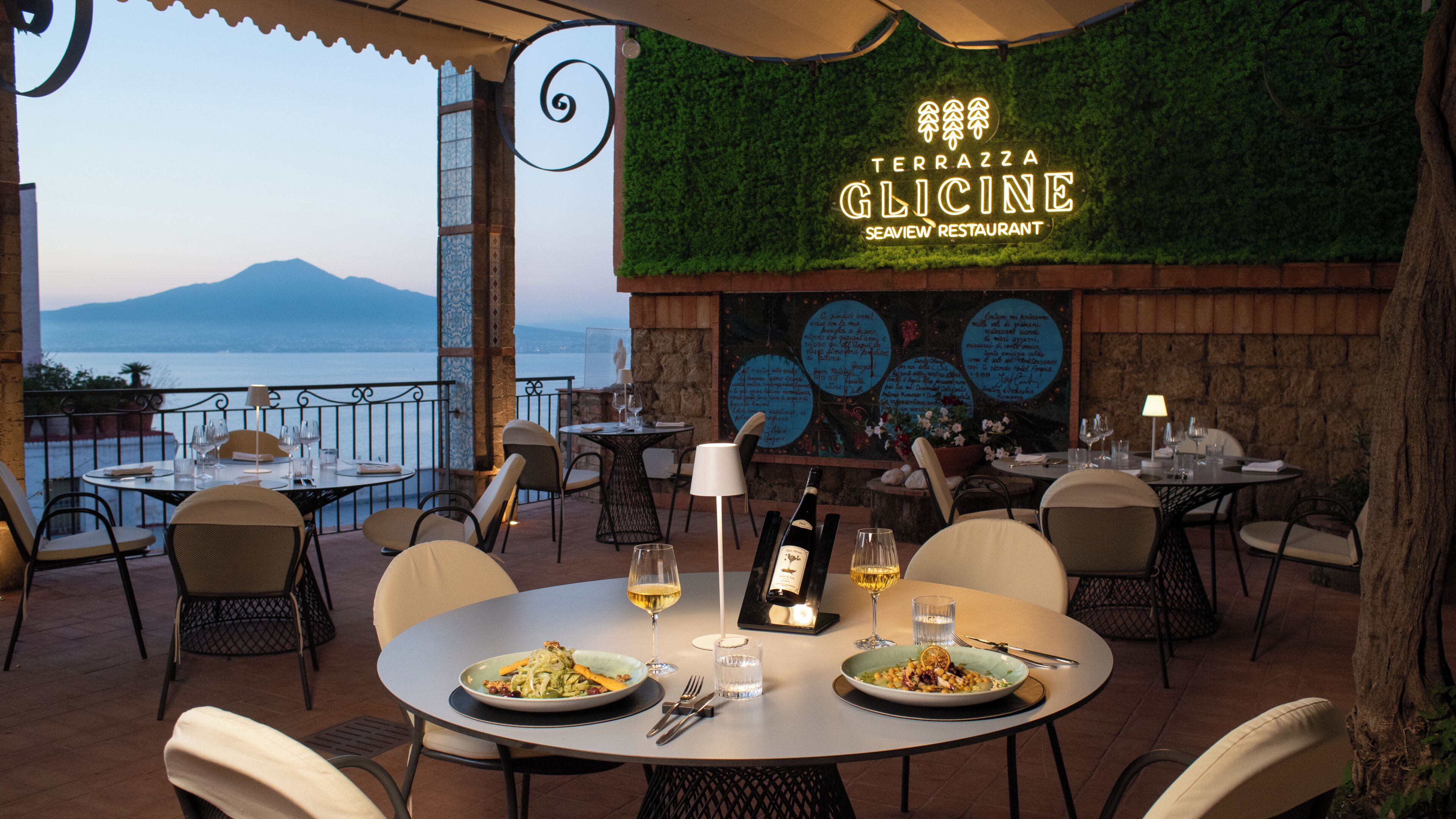 An outdoor restaurant with tables and chairs, a view of the sea, and a sign for 'Terrazza Glicine.'