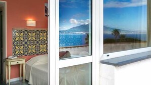 A hotel room with a bed, a nightstand, a lamp, and a window with a view of a mountain and the sky. - Hotel Aequa (Vico Equense)