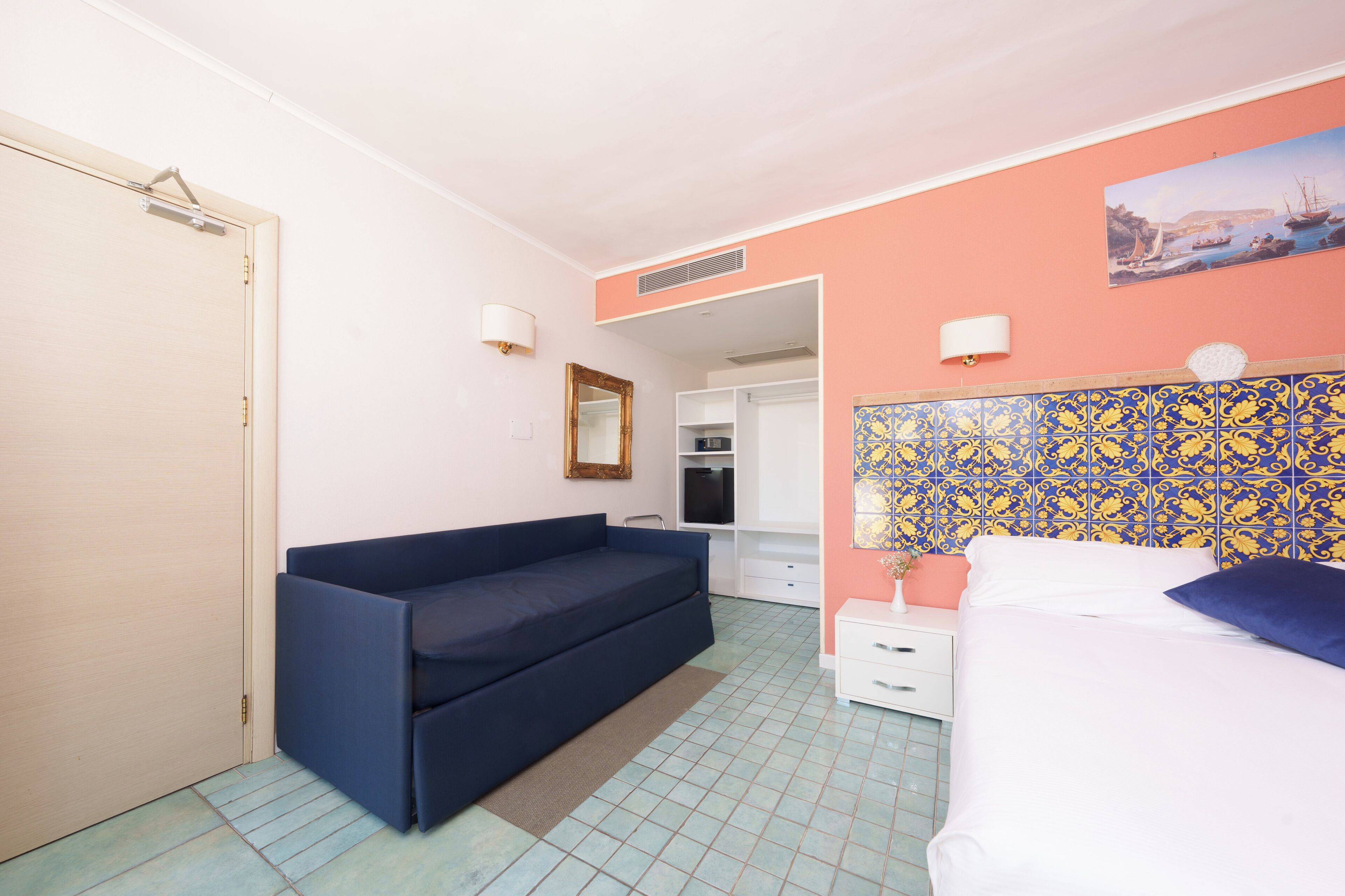 A bedroom with a blue sofa, a bed, a white nightstand, a mirror, and a tiled wall with a coastal painting.