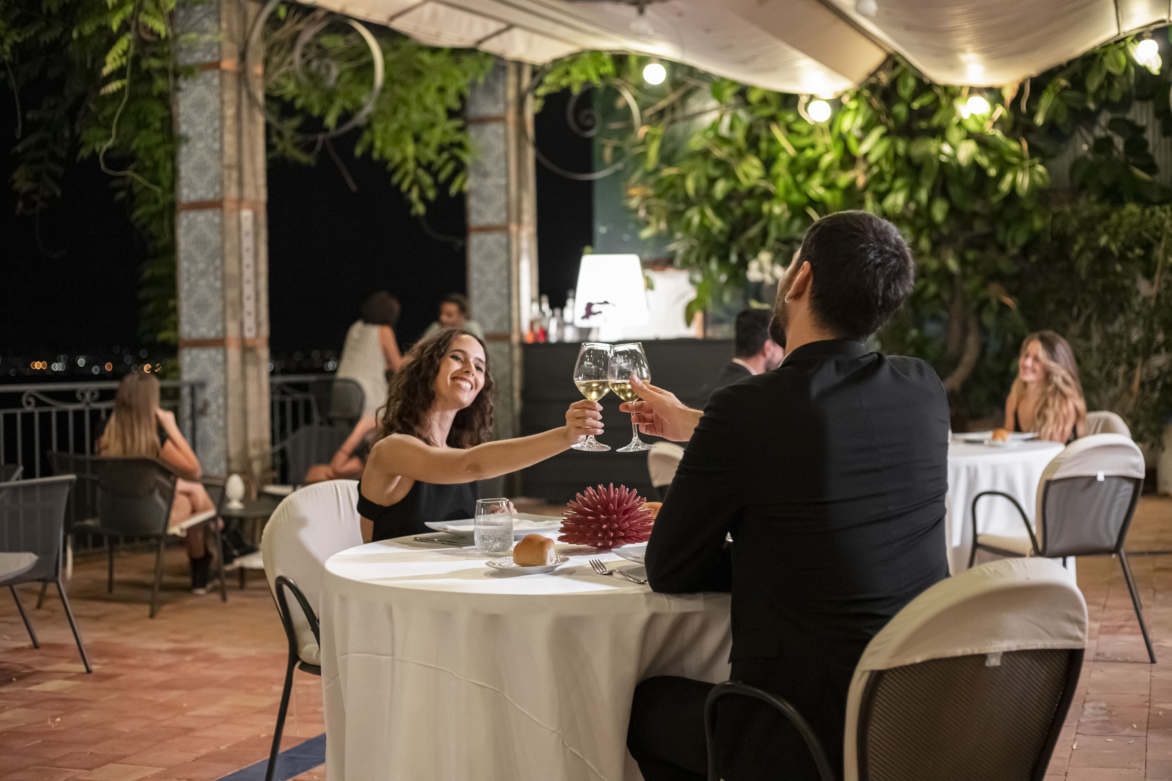 A couple toasting with wine glasses at an outdoor table with a white tablecloth, surrounded by chairs and a potted plant.