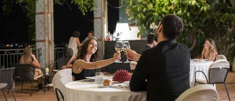 A couple toasting with wine glasses at an outdoor table with a white tablecloth, surrounded by chairs and a potted plant.
