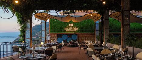 An outdoor restaurant with tables and chairs, a view of the sea, and a sign for 'Terrazza Glicine.'