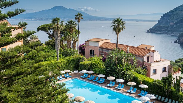 A pool area with lounge chairs and umbrellas, surrounded by greenery and overlooking a body of water with mountains in the background. - Hotel Aequa (Vico Equense)