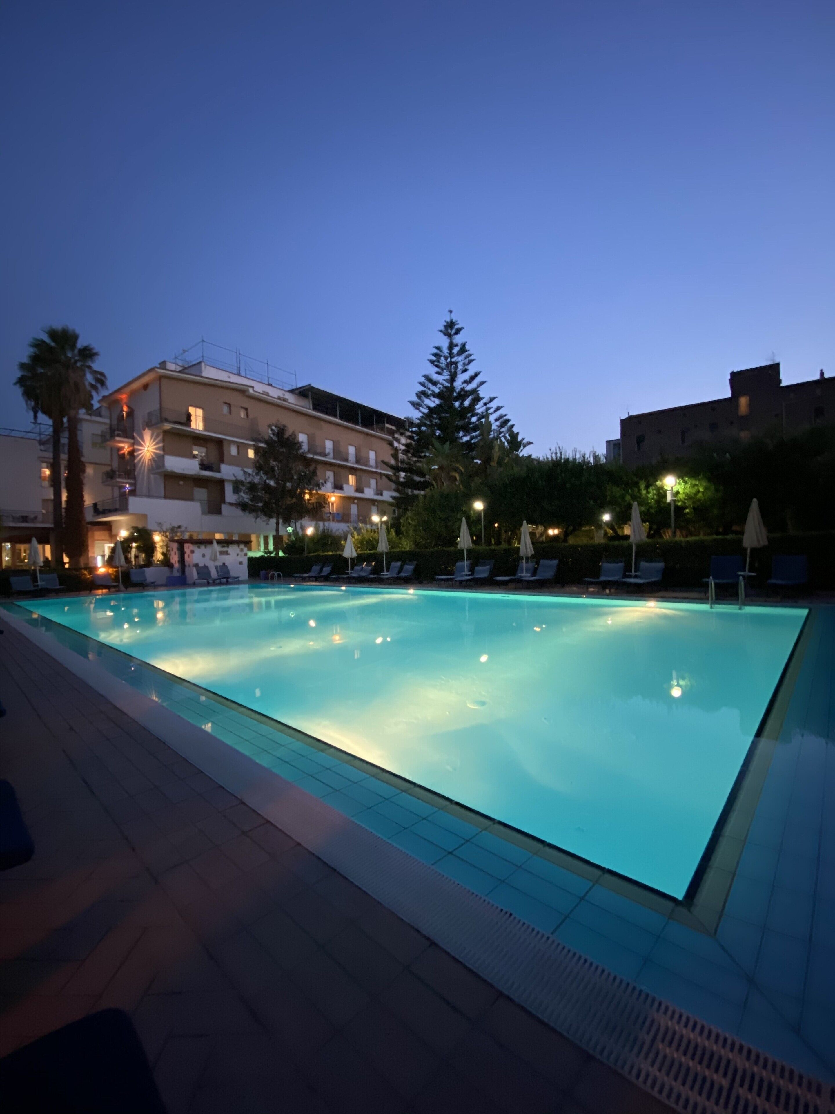 A swimming pool with lounge chairs and umbrellas, surrounded by buildings with balconies and lights.