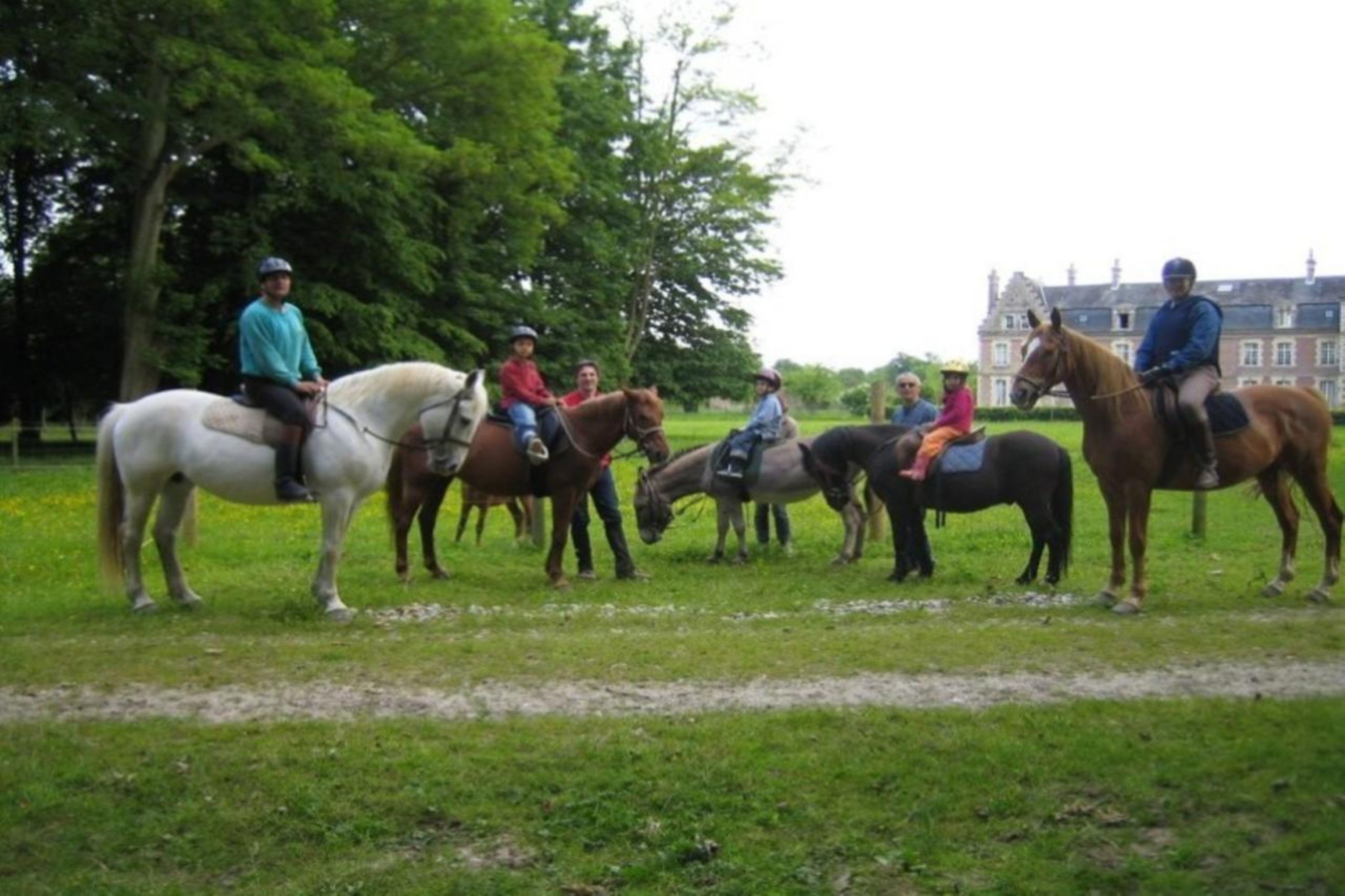 Um grupo de pessoas cavalgando em uma área gramada com árvores e um edifício ao fundo.