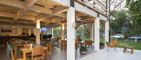 A spacious outdoor dining area with wooden tables and chairs, a bar area, and a view of a garden and parked cars.